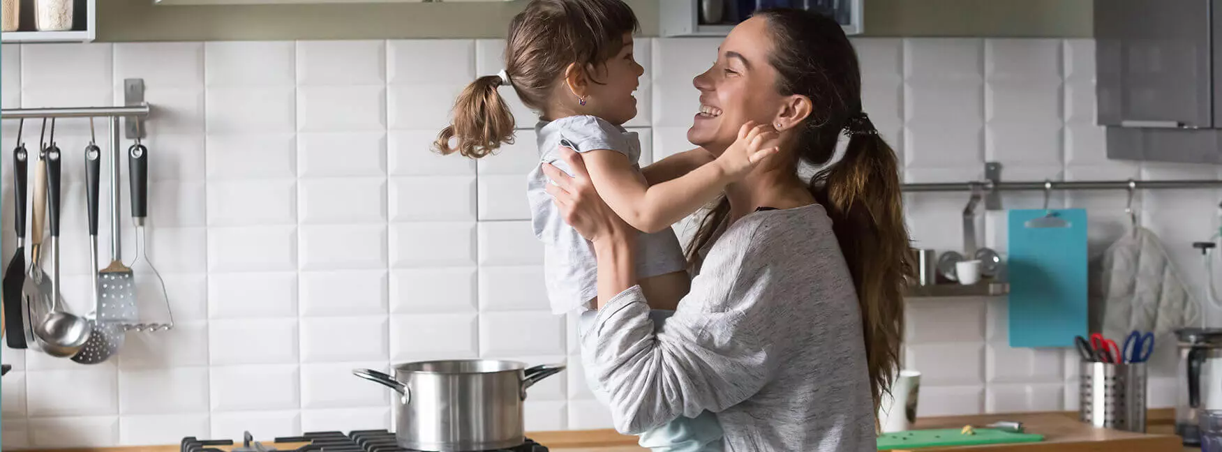 A parent in the kitchen holding their child up in their arms while they smile at one another.